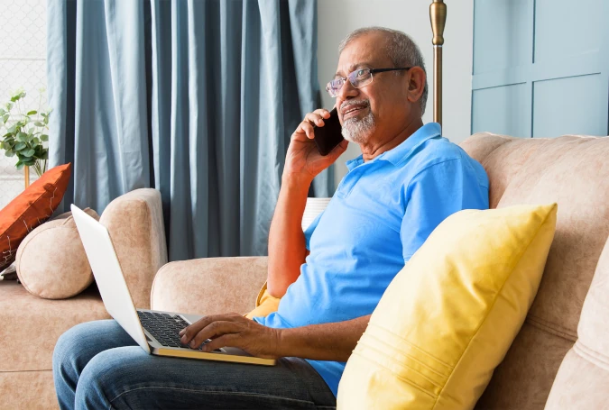 a man in a blue shirt sitting on a sofa, using a laptop while speaking on a telephone